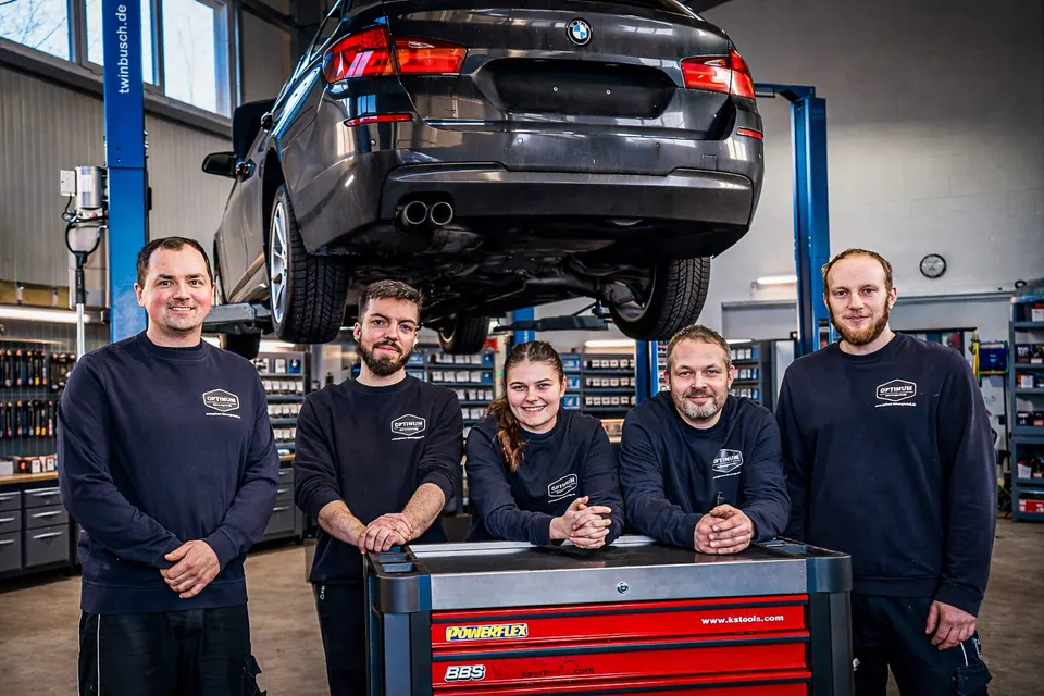 Das Optimum Fahrzeugtechnik Team mit 5 Experten in schwarzen Shirts vor einem BMW auf der Hebebühne in der Werkstatt
